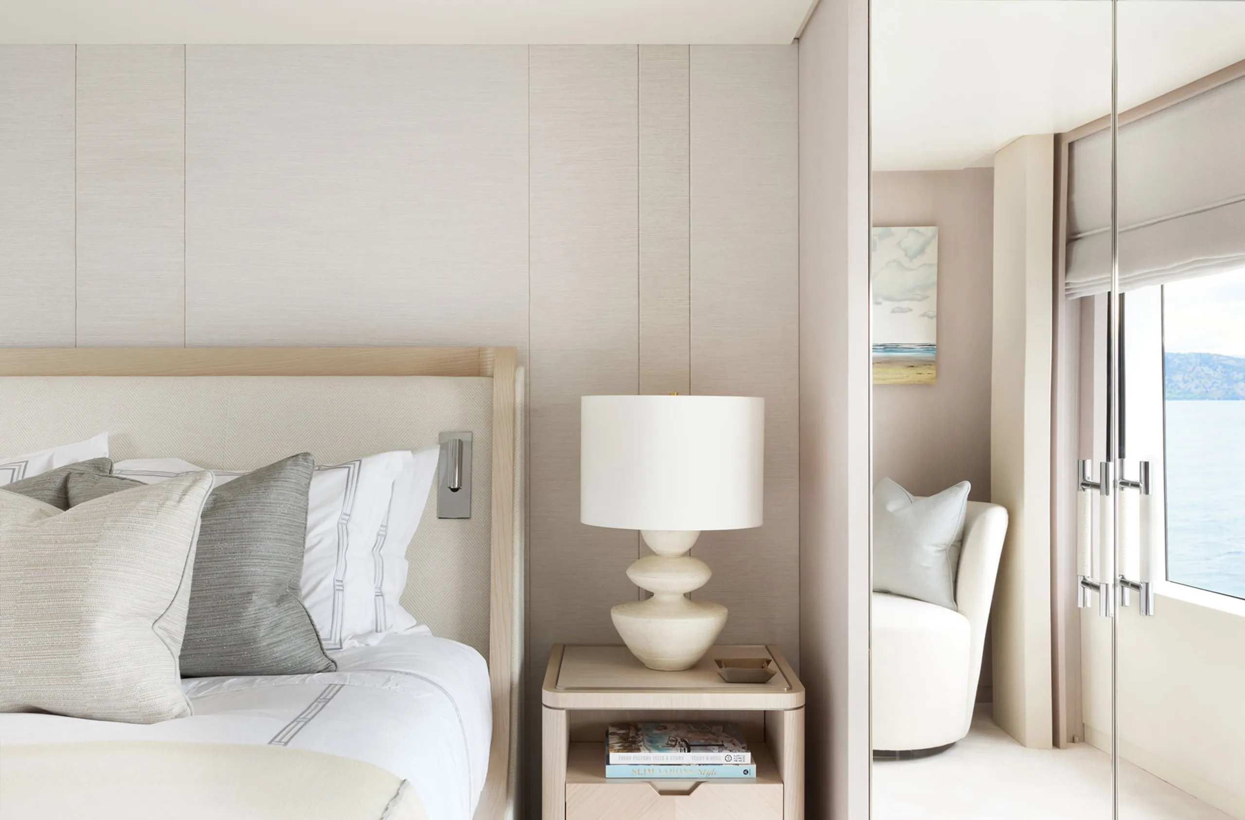 Mid shot details of the main suite on the superyacht, a mirror is reflecting a small arm chair on the right third of the image, in the middle is a bedside table with decorative bespoke lamp and the left third showcases a part of the bed